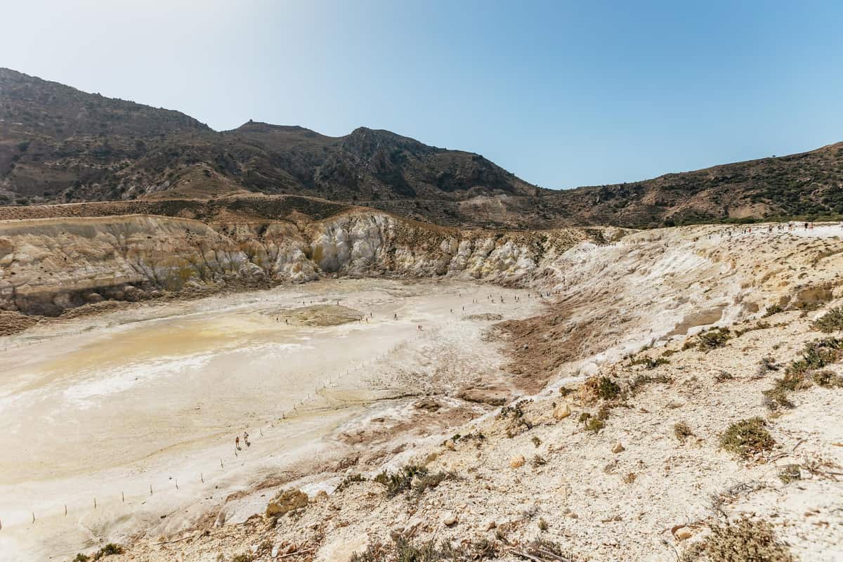 Nisyros volcano tour visitors walking across the pale floor of Stefanos crater, seen from the rim under a blue sky