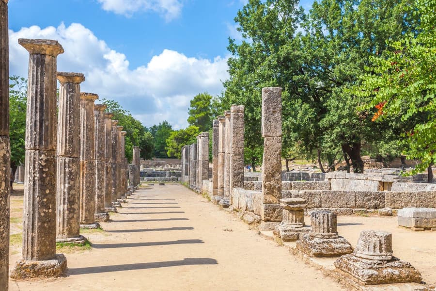 Olympia day trip: rows of tall fluted stone columns line a sunlit sandy path through ancient ruins, with green trees and a blue sky with white clouds.