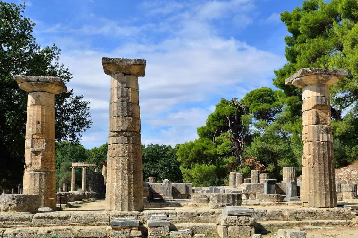 Olympia day trip: three tall weathered stone columns of an ancient temple stand on a stone platform under a blue sky with white clouds, with pine trees and green hills behind.