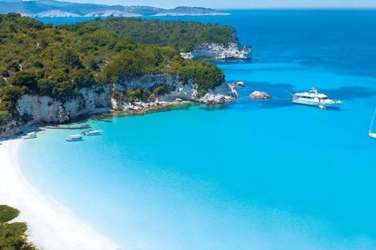 Paxos beach trip: an aerial view of a turquoise bay with a white sand beach curving along a wooded green headland, with a white boat and a sailing yacht anchored on the clear blue water.
