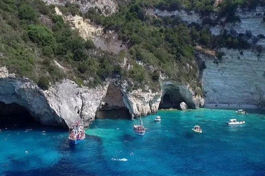 Paxos beach trip: white limestone cliffs with sea arches and a dark cave rise above turquoise water, with small boats and swimmers on the water below green hills.