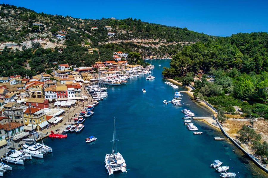 Paxos boat trip: an aerial view of a narrow blue harbour inlet with moored sailing yachts and small boats along a waterfront of orange-roofed houses, with green wooded hills under a deep blue sky.