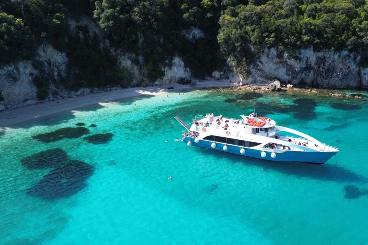 Paxos boat trip: a white and blue passenger boat with people on its decks is anchored in clear turquoise shallow water beside a pebble beach below tall wooded cliffs, with a few swimmers in the water.