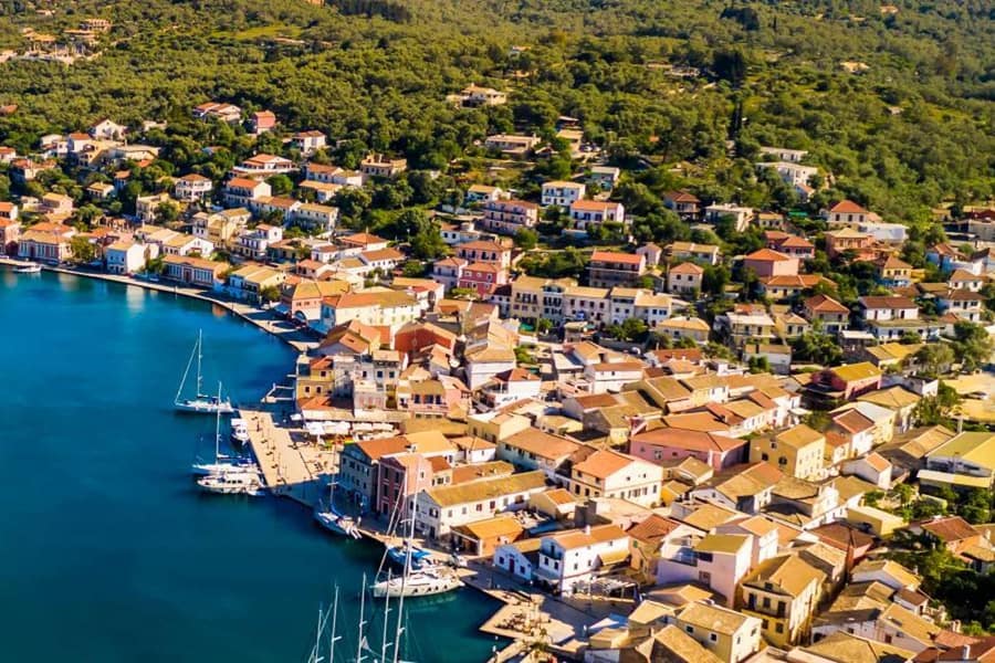 Paxos day trip: an aerial view of a small harbour town with pastel houses and orange-tiled roofs along a stone quay, sailing boats moored in the deep blue water and green wooded hills behind.