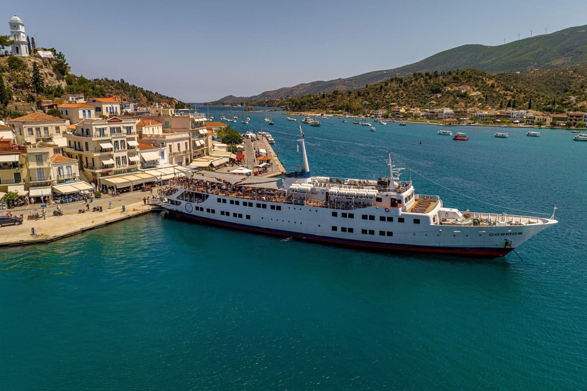 Poros boat trip: a white cruise ship with passengers on its open decks lies alongside the quay of a waterfront town, with a white clock tower on the hill, pastel houses with terracotta roofs, calm turquoise water and small boats anchored across the strait below green hills.