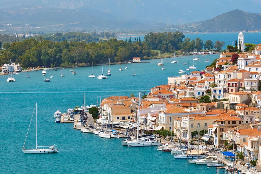 Poros boat trip: a hillside town of white and terracotta-roofed houses runs down to a quay lined with moored sailing yachts, with a white clock tower on the ridge, anchored sailboats on the turquoise strait and wooded green hills across the water.