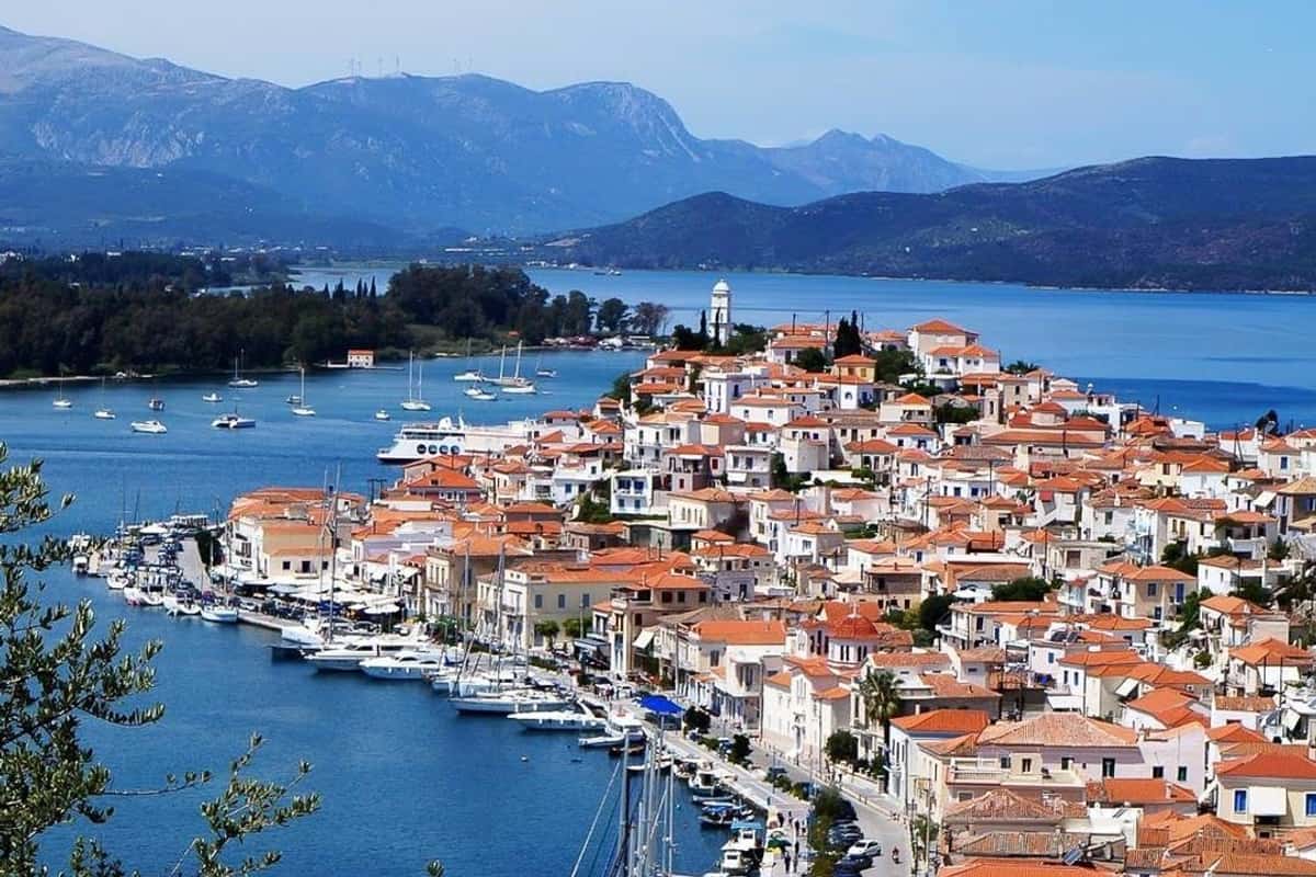Poros day trip: a view from a hillside, past a leafy branch, over a waterfront town of white and terracotta-roofed houses on a peninsula, with a white clock tower at its far end, a harbour lined with moored yachts and sailing boats, a calm blue strait and layers of mountains behind under a pale sky.