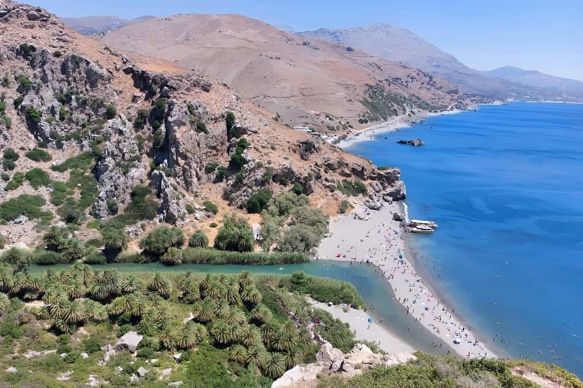 Preveli beach tour: Preveli Palm Beach seen from a clifftop viewpoint, with a green palm grove and a turquoise river meeting a long pale-sand beach where swimmers and two tour boats sit beside deep blue sea, brown mountains rising behind under a clear sky.