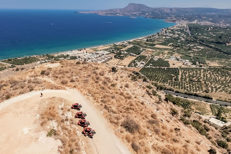 Rethymno ATV tour: an aerial view from a dry hilltop of four red buggies parked on a pale dirt track above a long bay, with deep blue sea, white buildings along the shore, green groves, a road and a mountain cape in the distance.