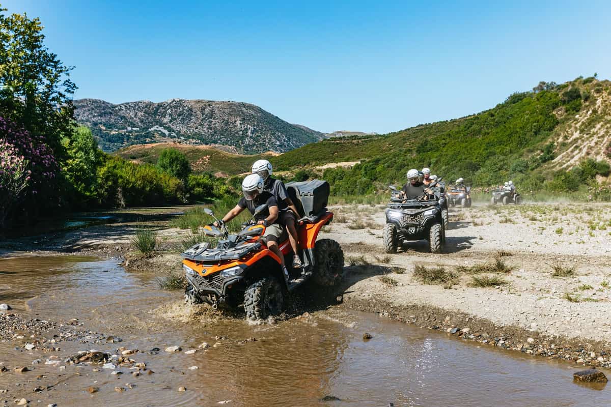 Rethymno ATV tour: an orange quad bike with two helmeted riders splashes through a shallow brown stream beside a gravel bank, with more quad bikes following in a line and green hills and a rocky mountain behind them under a clear blue sky.
