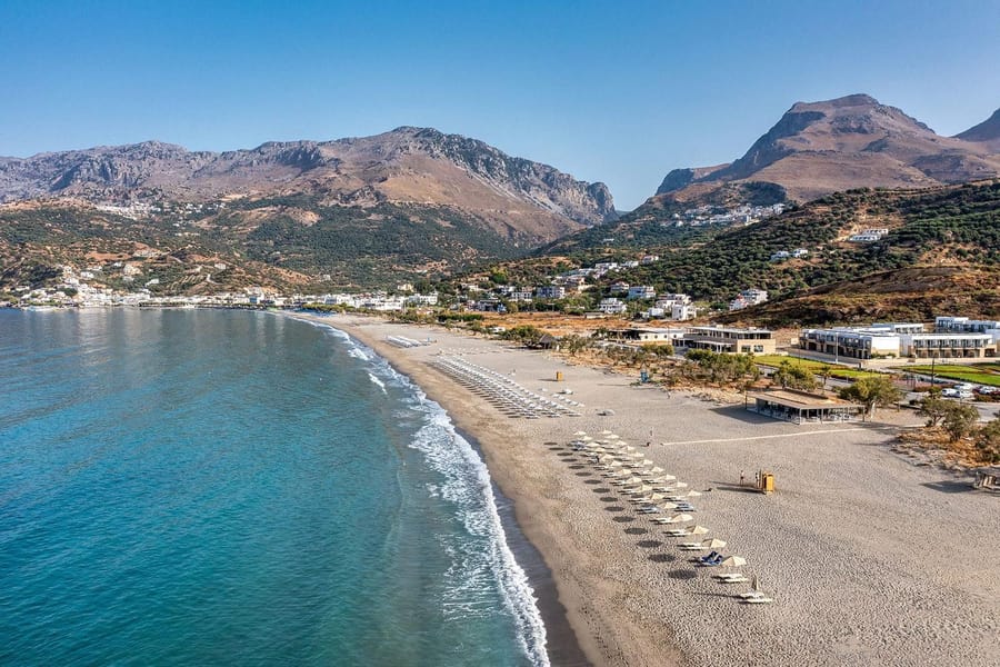 Rethymno beach trip: an aerial view along a long curving sandy beach with rows of parasols beside calm turquoise water and white surf, a white village at the foot of rugged brown mountains and a clear blue sky.
