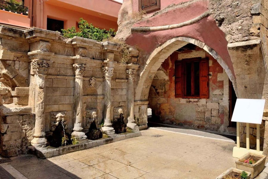Rethymno day trip: a weathered stone fountain wall with slender columns, carved capitals and lion-head spouts stands beside a pointed stone arch with a red-shuttered window on a sunlit paved square.