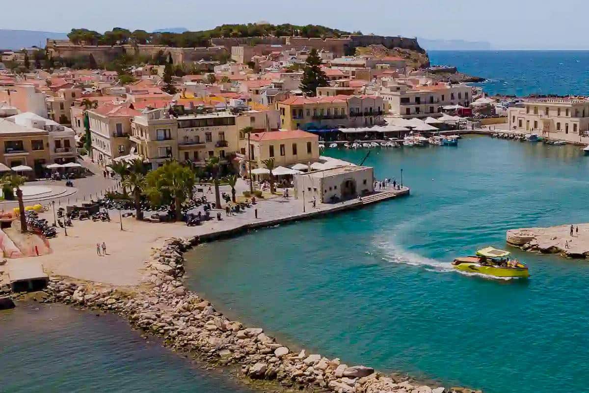 Rethymno tickets: an aerial view of old town houses with terracotta roofs and palm trees beside a curved stone quay and a turquoise harbour, with a yellow tour boat crossing the water and a long fortress wall on a pine-covered hill behind the town under a pale blue sky.