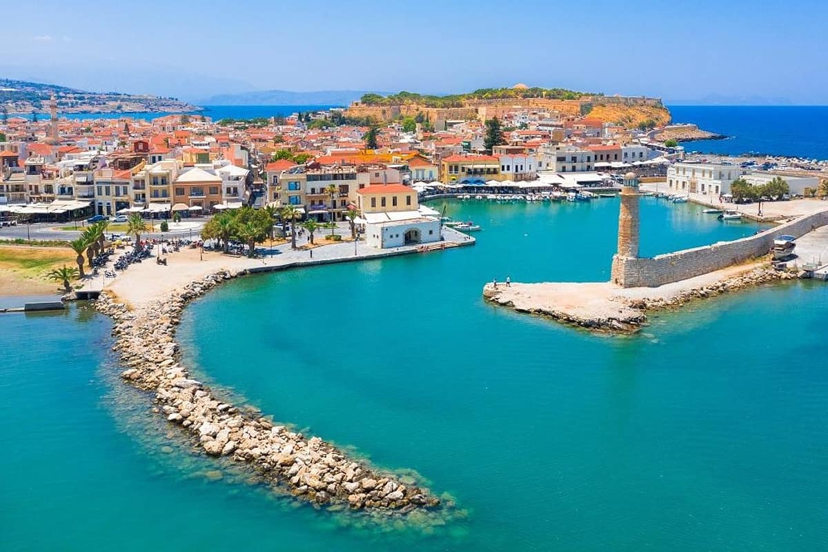 Rethymno transfer: an aerial view of the Venetian Harbour of Rethymno with its old stone lighthouse at the end of the breakwater, turquoise water, waterfront cafes and old town houses with terracotta roofs, and the Fortezza fortress walls on the hill behind under a clear blue sky.
