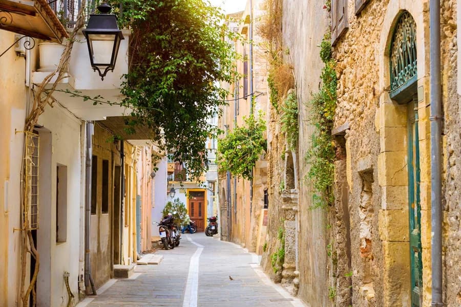 Rethymno transfer: a narrow sunlit old town lane between cream and golden stone houses, with a black street lantern, green climbing plants over a balcony, a green arched door on the right and two parked scooters at the far end.