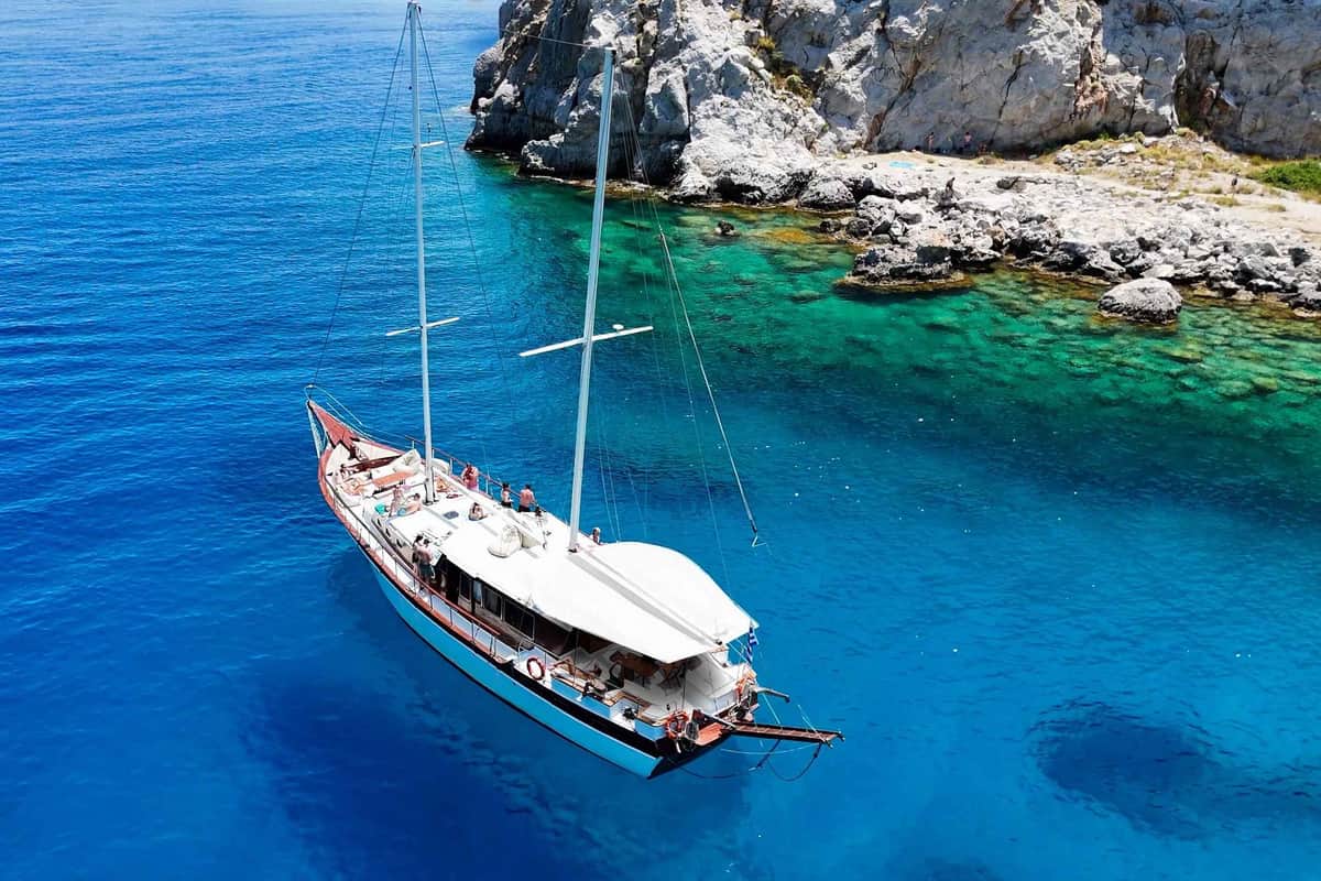 Rhodes boat trip: a white two-masted wooden sailing boat with people on deck lies at anchor on deep blue water beside a rocky shore with clear green shallows.