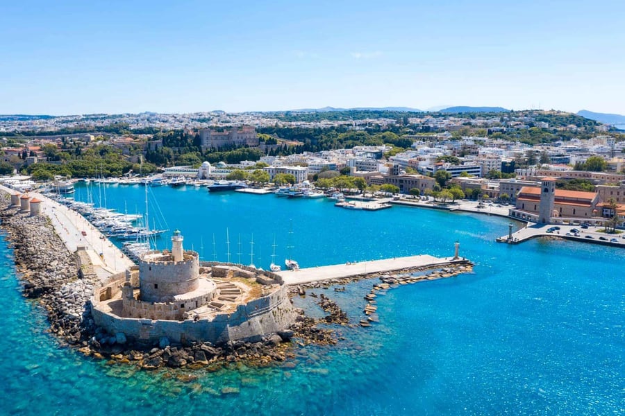 Rhodes day trip: an aerial view of a harbour with a round stone fort and lighthouse at the end of a breakwater, a row of windmills on the mole, moored yachts, a bell tower on the quay and the town spreading behind under a clear blue sky.
