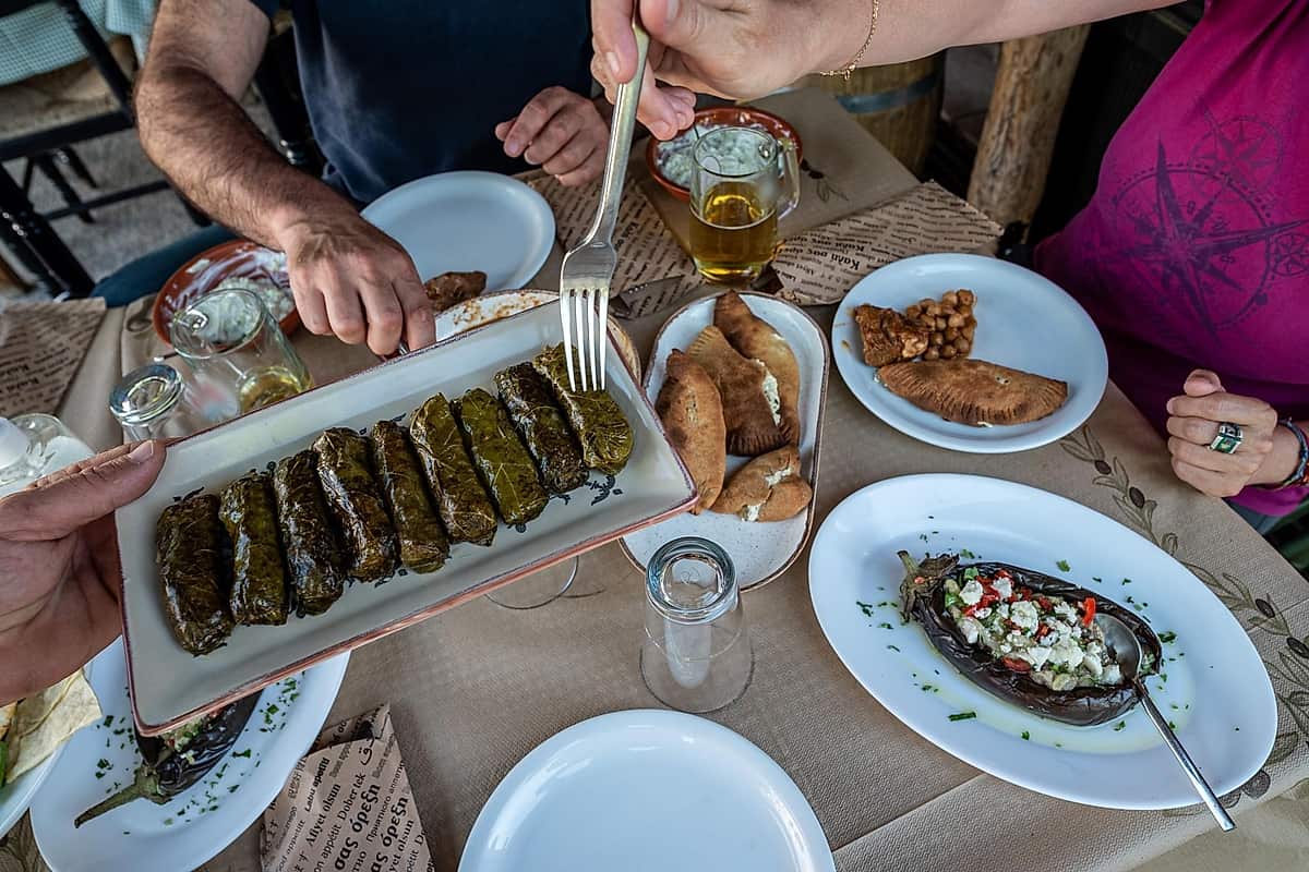 Rhodes food tour: an overhead view of a restaurant table with a platter of stuffed vine leaves held by one hand, a plate of golden half-moon pastries, a roasted aubergine topped with feta, chopped tomato and herbs, a plate of chickpeas and a glass of beer, with a fork reaching in from above.