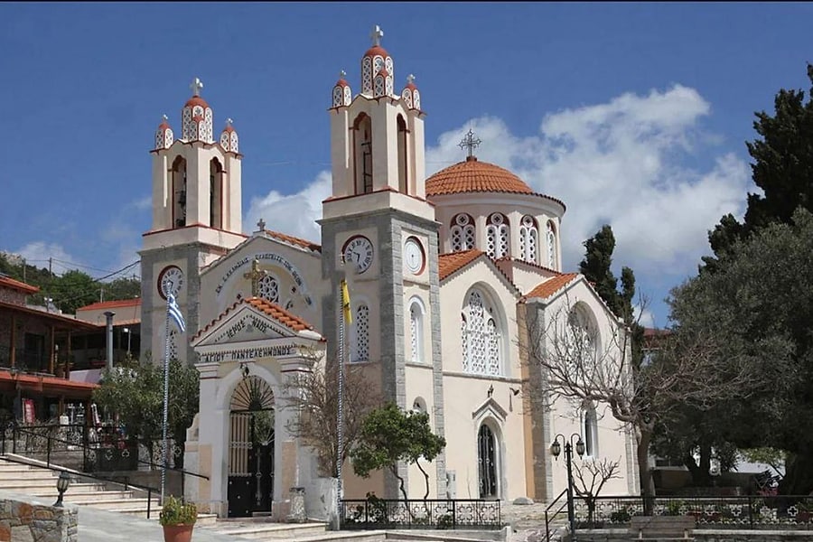 Rhodes food tour: a cream-coloured church with two bell towers topped with small red-and-white domes and a terracotta-tiled central dome stands beside a paved village square and green trees under a deep blue sky with white clouds.