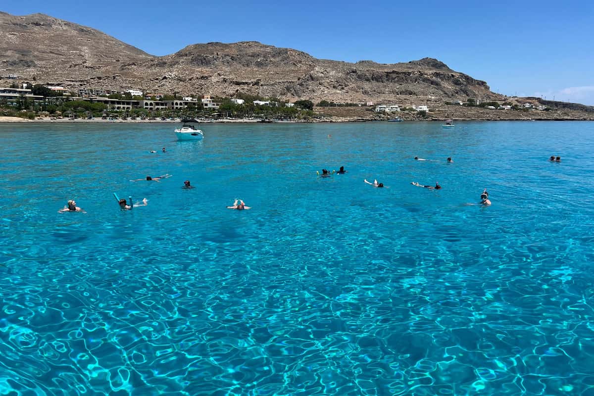 Rhodes snorkeling tour: a wide bay of clear turquoise water with rippling light patterns, a few snorkelers floating far from the camera and a white boat at anchor, with a sandy shore, low buildings and dry brown hills under a blue sky.