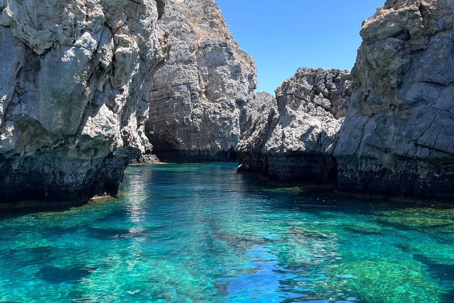 Rhodes snorkeling tour: a narrow rocky inlet between tall grey limestone cliffs, with clear turquoise and green water over the sea floor under a bright blue sky.