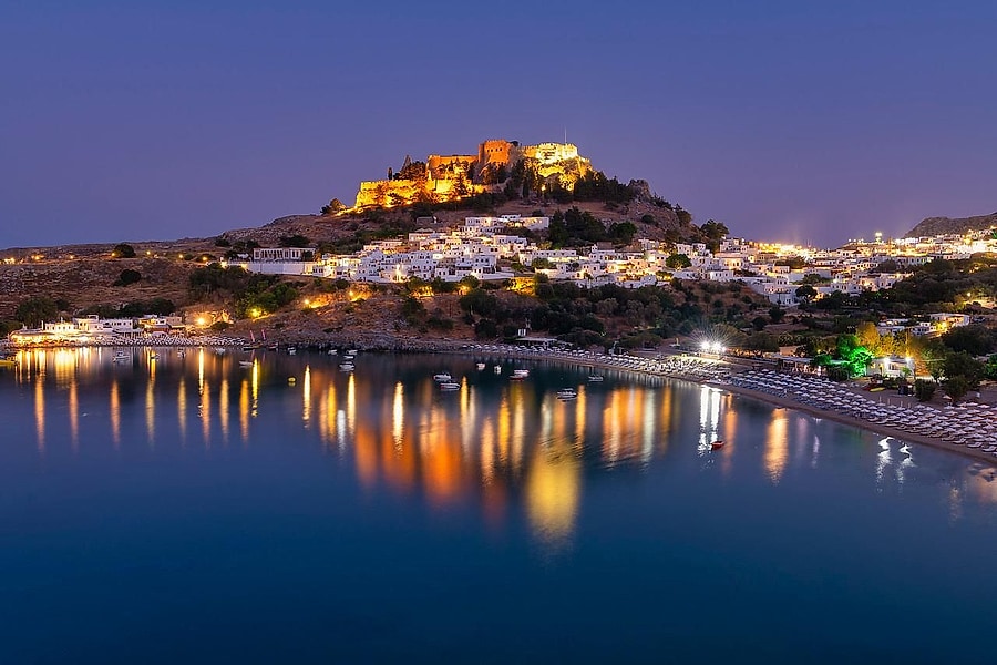 Rhodes sunset cruise: the floodlit Acropolis of Lindos glows gold on its hilltop above the white houses of Lindos village, with the lights of the bay reflected in the calm dark-blue water and rows of sunshades along the beach under a purple evening sky.