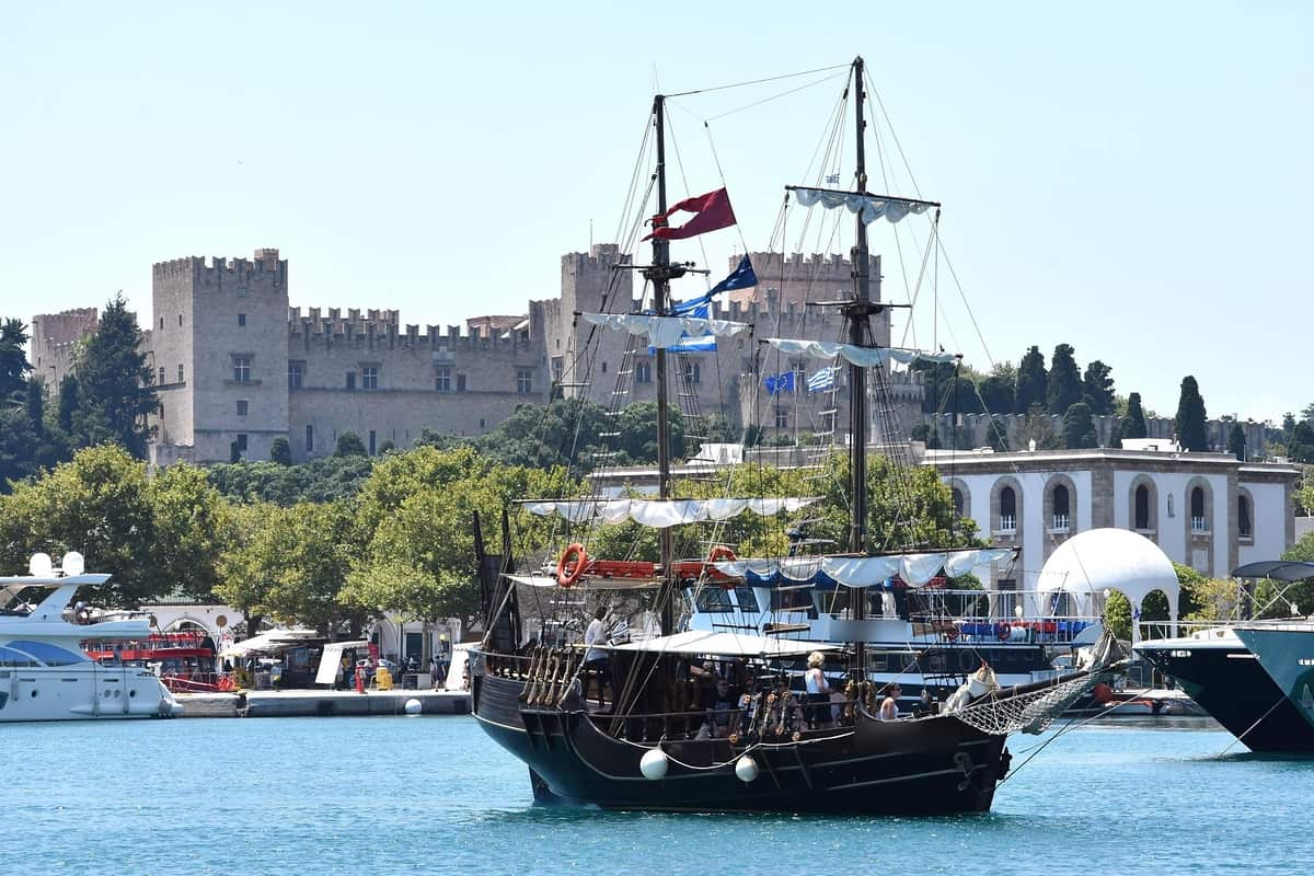 Rhodes sunset cruise: a dark wooden two-masted sailing ship with white sails and Greek flags floats in the harbour of Rhodes Town in front of the crenellated towers of the Palace of the Grand Master, with white motor yachts moored beside it under a clear blue sky.