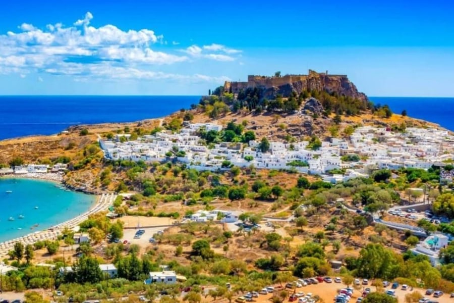 Rhodes transfer: a rocky hill crowned by a stone fortress rises above a village of white houses, with a turquoise bay and sandy beach on the left, green trees below and a deep blue sea behind.