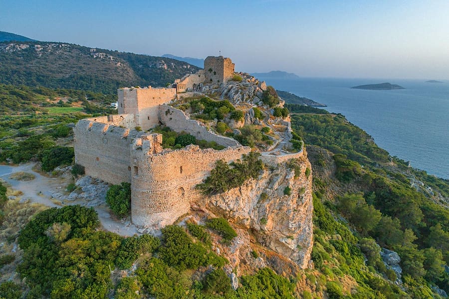 Rhodes wine tour: an aerial view of a stone castle ruin with a square tower on a rocky hilltop, with pine-covered green hills and a calm blue sea with small islands behind.