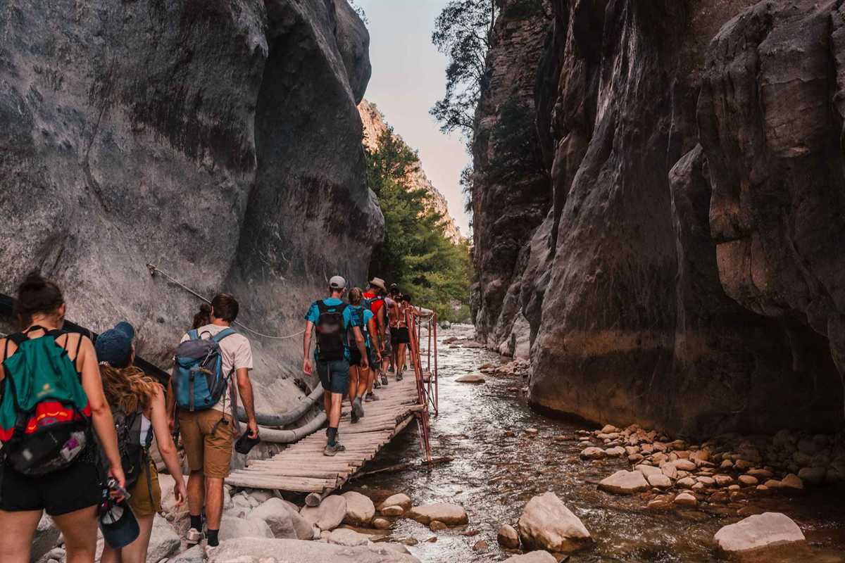 Samaria gorge tour: a line of hikers with backpacks walks along a wooden plank walkway beside a stream between tall grey rock walls.