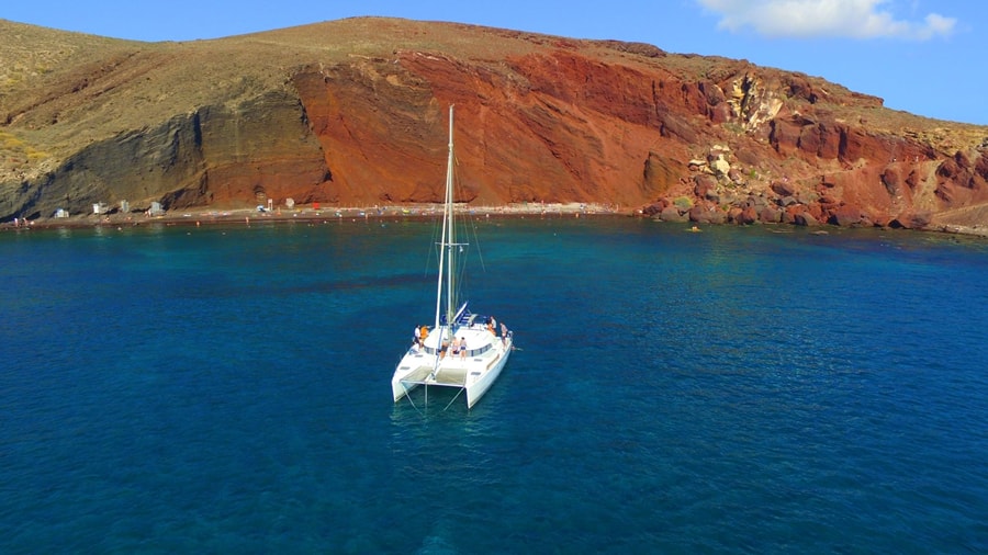 Santorini catamaran cruise anchored in blue water off Red Beach, with the red volcanic cliffs behind