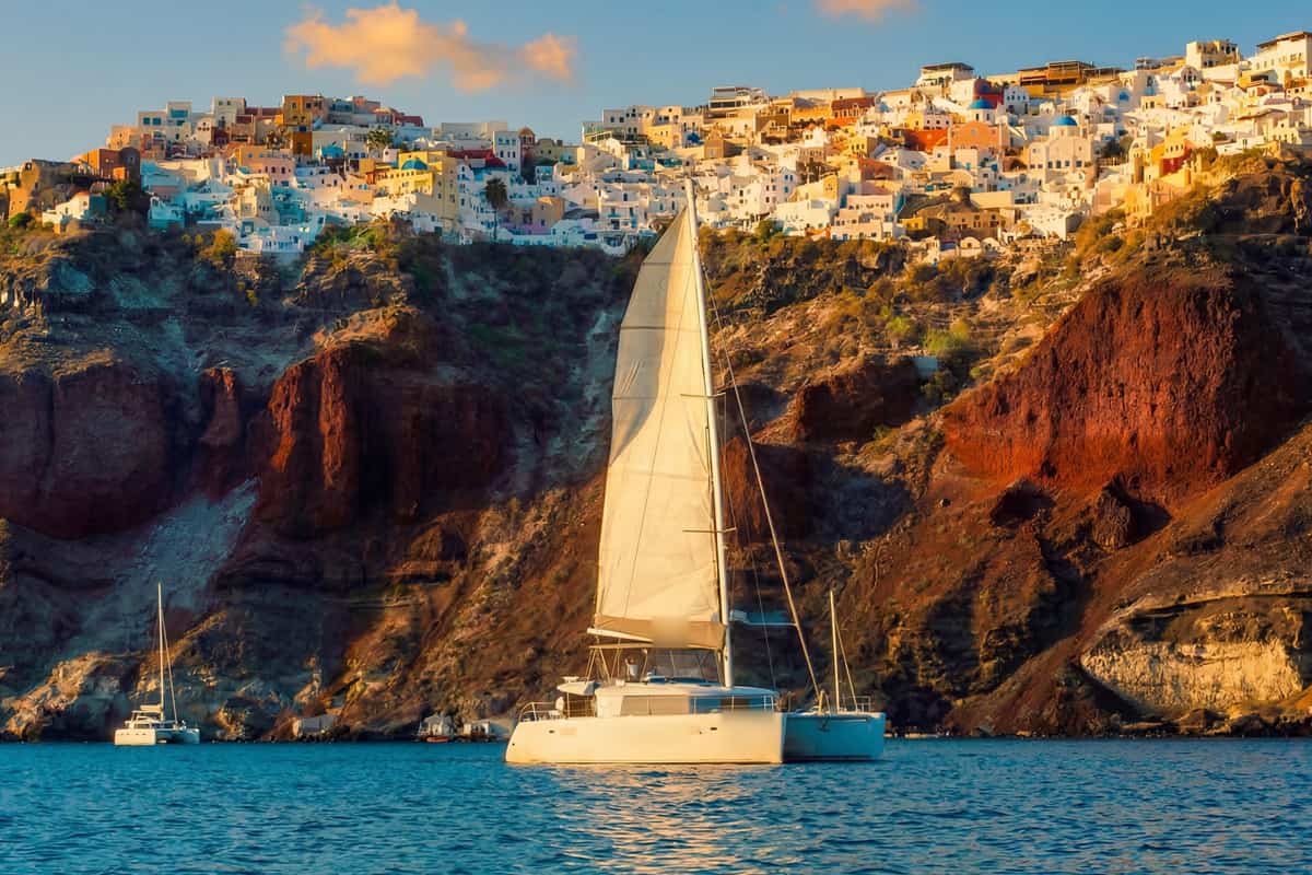 Santorini catamaran cruise under sail below the red cliffs and white houses of Oia in golden light