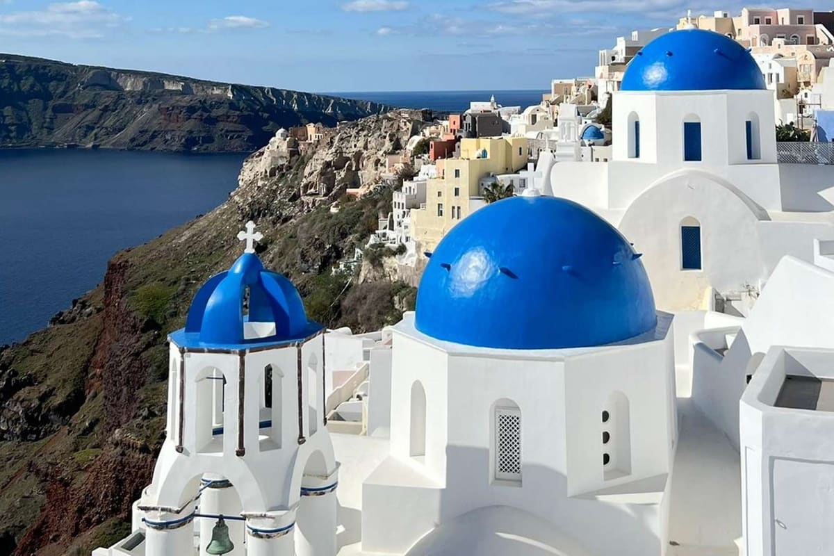 Santorini day trip: two bright blue church domes and a white bell tower with a green cross stand among white and pastel-colored houses on a steep cliff above deep blue water, with a dark green cliff across the bay under a blue sky.