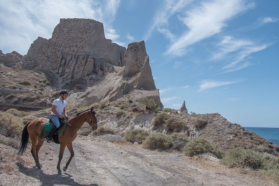 Santorini horse riding: a brown horse with a rider in a white shirt and helmet walks along a sandy track below a tall pale rock formation full of small holes, with a strip of blue sea on the right under a bright blue sky with wispy clouds.