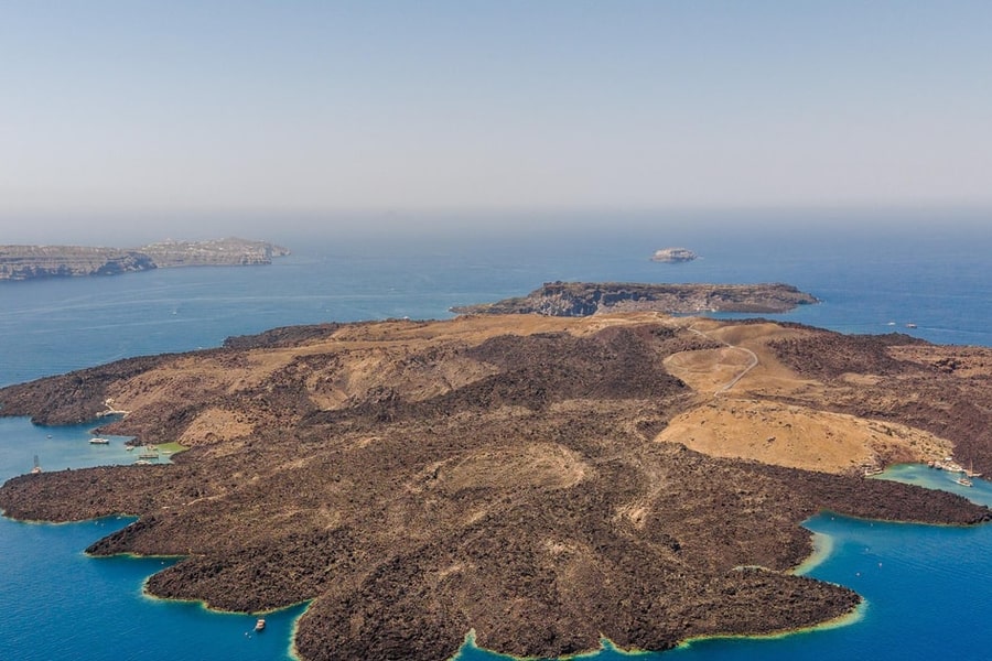 Santorini spa: aerial view of a dark, barren volcanic islet with a pale track winding across its top, blue sea around it and cliffs on the far side under a pale sky.