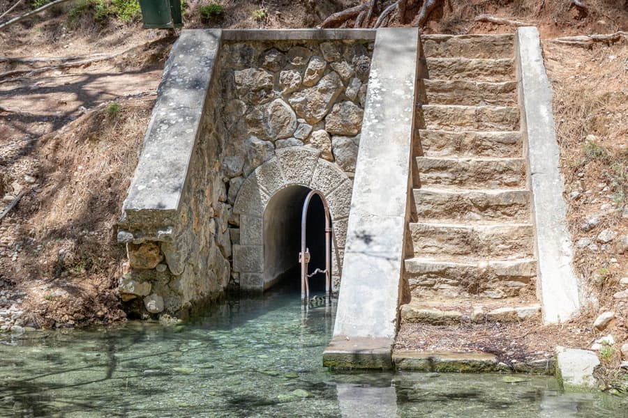 Seven Springs Rhodes: the arched stone mouth of the Epta Piges spring tunnel with an iron gate, a wide stone staircase beside it and clear shallow spring water in the foreground.