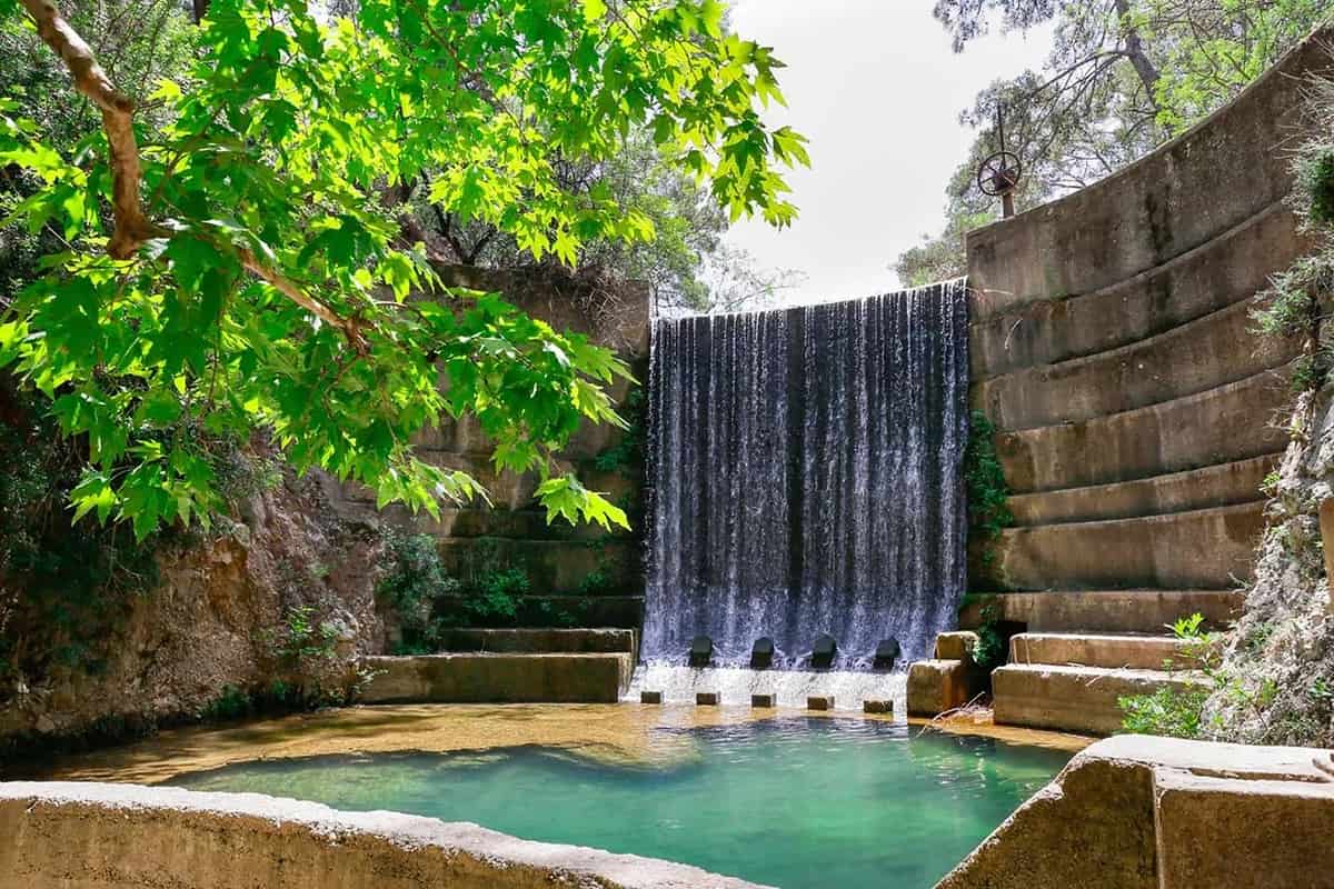 Seven Springs Rhodes: the dam at Epta Piges with a curtain of water falling into a turquoise pool, framed by sunlit green plane tree leaves, curved stone steps and tall trees.