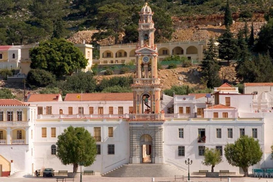 Symi boat trip: a whitewashed monastery with a tall, ornate clock and bell tower rising above a stone gateway and wide steps, with pine trees on the hill behind.