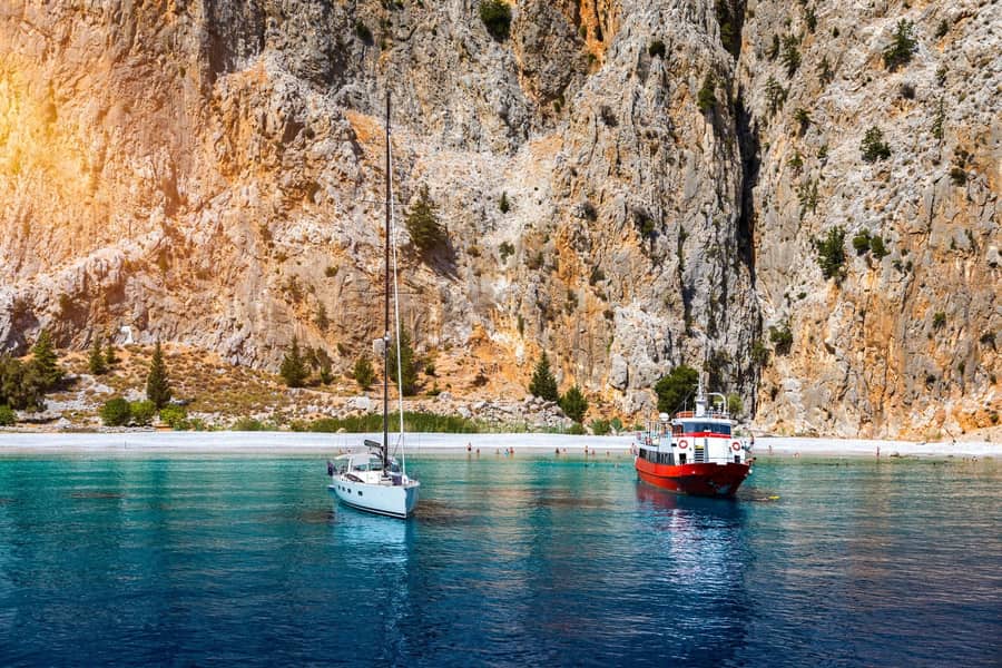 Symi day trip: a tall cliff of grey and orange rock rises above a long pale pebble beach, with a white sailboat and a red and white boat anchored on calm turquoise-blue water in front of it.