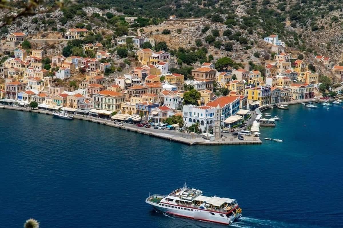 Symi day trip: a high view of Symi harbour, with rows of pastel yellow, orange and white houses climbing the green hillside above a quay lined with cafes, a slim stone clock tower at the end of the quay, and a white passenger boat crossing the deep blue water in the foreground.
