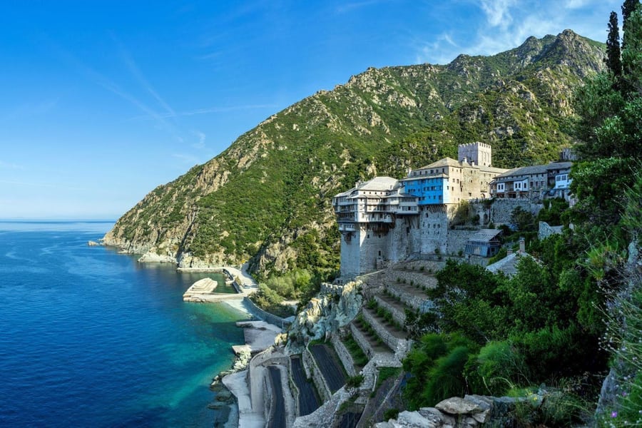Thessaloniki day trip: a large stone monastery with white and blue wooden balconies and a square tower stands on a terraced shore above deep blue sea, with a steep green mountain slope behind it under a bright blue sky.