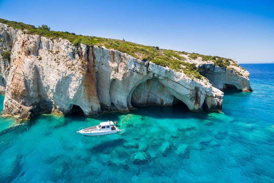 Zakynthos beach trip: a tall white limestone sea cliff topped with green scrub has large arched caves at the waterline above clear turquoise water, with a small white motorboat floating below the cliff under a deep blue sky.