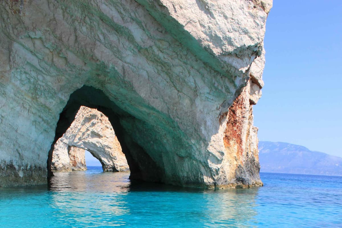 Zakynthos blue caves tour: a tall white limestone cliff streaked with rust-coloured rock has two large sea arches that open onto bright turquoise water, with a distant mountain coast on the horizon under a clear blue sky.