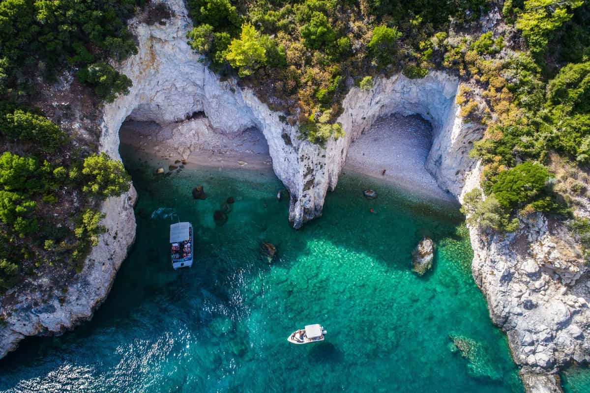 Zakynthos boat trip: an aerial view of a tour boat with a pale canopy and a small white motorboat on clear green-turquoise water in a cove below white limestone cliffs with two shallow caves, pebble shores and green scrub.