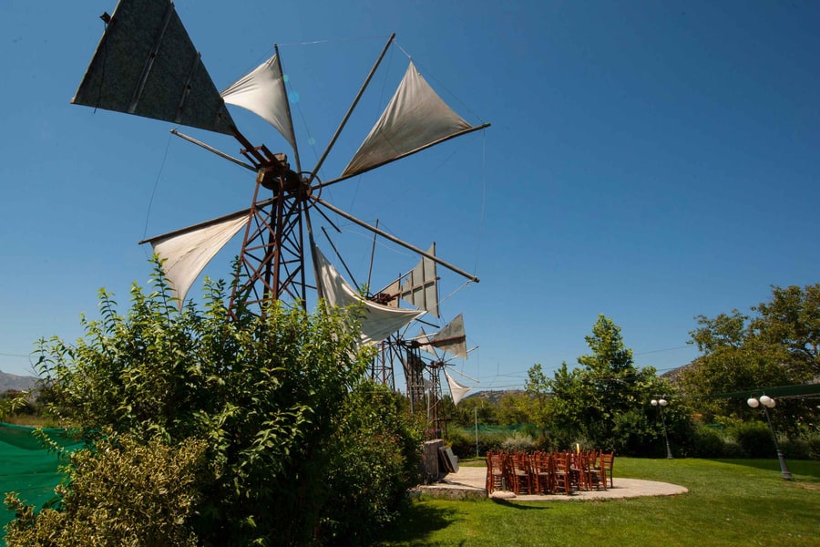 Zeus cave tour: a tall old windmill with white cloth sails stands against a deep blue sky beside green bushes, with more windmills behind it, a green lawn and wooden chairs on a stone patio.